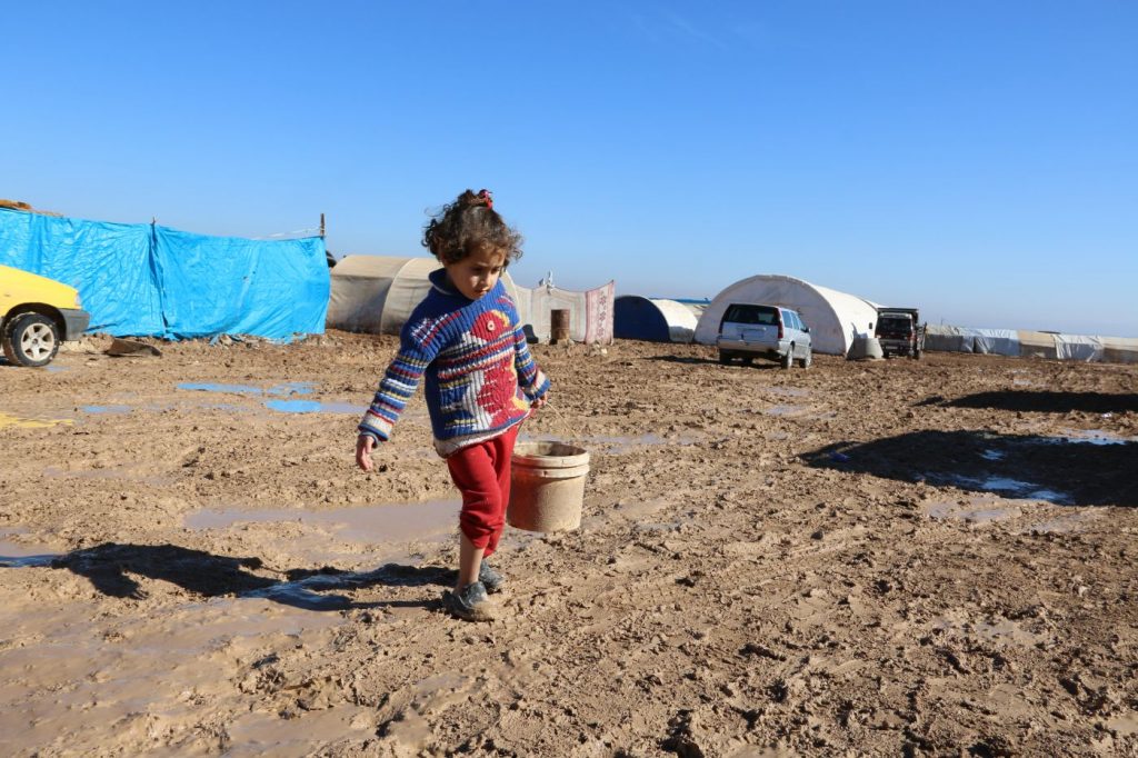 a young girl carrying a bucket through a refugee camp